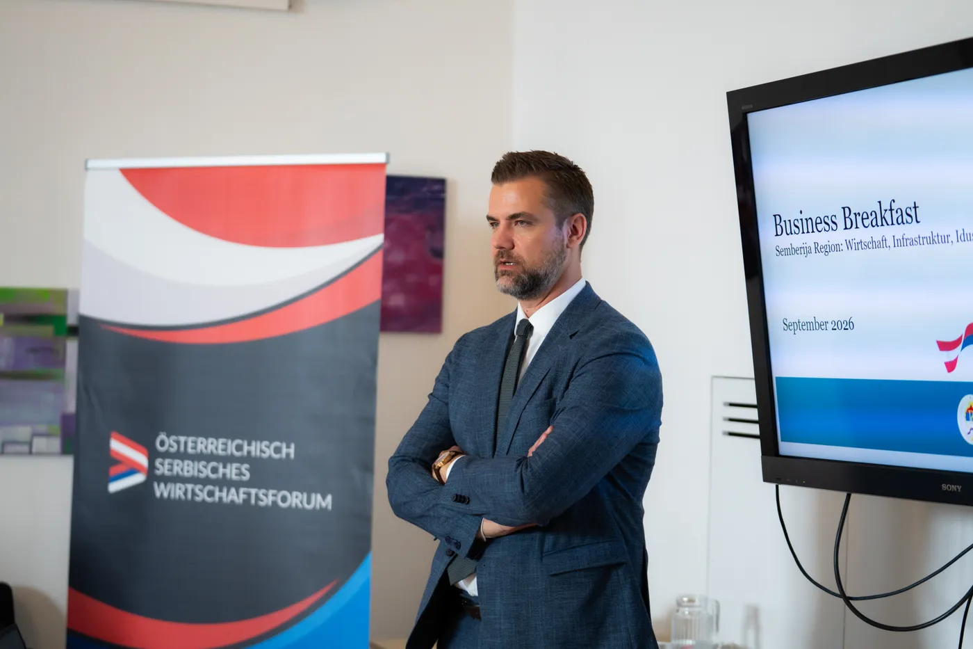 Vortragender vor der Titelfolie des Business Breakfast, daneben der Aufsteller des Wirtschaftsforums