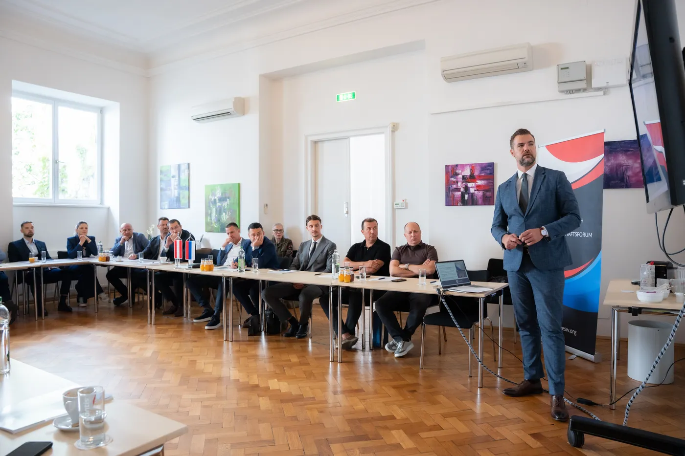 Blick in den Saal: der Vortragende vor dem Aufsteller, die Teilnehmer an der langen Tafel